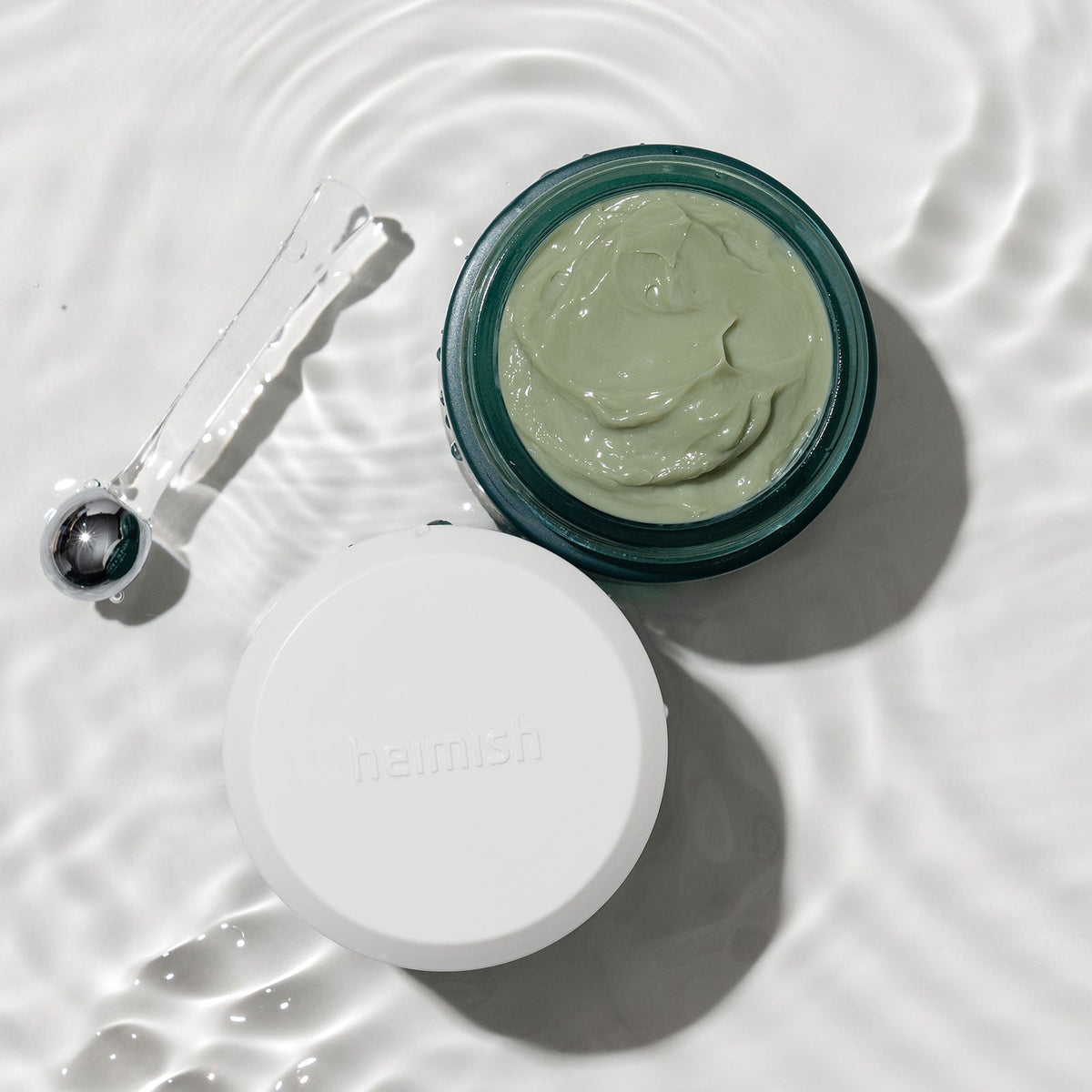 Heimish Marine Care Eye Cream - 30ml — overhead view of the open eye-cream jar, lid and metal applicator on a water-textured surface.